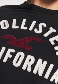 Camiseta negra con letras en relieve blancas que dicen "Hollister California" y un logo de gaviota bordado en rojo, sobre una tela lisa.