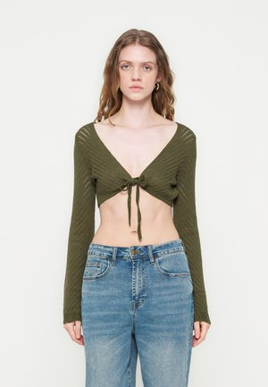 Frau trägt einen olivgrünen, gestrickten cropped Pullover mit Knotendetail vorne, langen Ärmeln und hoch taillierte blaue Jeans vor einfarbigem Hintergrund.