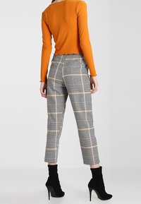 Frau in orangefarbener langärmeliger Bluse, hoch taillierten karierten Hosen und schwarzen hochhackigen Stiefeletten, steht mit dem Rücken zur Betrachterin.