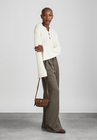 Femme portant un pantalon large gris, un haut à manches longues blanc à côtes, tenant un sac à bandoulière marron, se tenant debout sur un sol gris contre un fond uni.