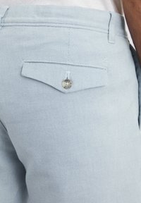 Pantalons chino bleu clair en tissu texturé. Comprend une poche arrière avec une fermeture à bouton et une petite étiquette au-dessus de la poche.