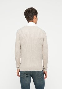 Lys beige strikket genser med ripet tekstur, lange ermer og rund hals, iført over en hvit kraged skjorte, sett fra baksiden.
