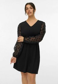 Zwarte jurk met een V-hals, korte lengte en een getailleerde taille. Beschikt over sheer, polka dot ballonmouwen van lichte stof.