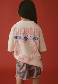 Kermankotoinen ylisuuri t-paita, jossa on vaaleanpunainen ja sininen "BLONDIE HEART of GLASS" -teksti sekä sydänkuvio. Yhdistetty vaaleanpunaisiin ja sinisiin raidallisiin shortseihin.