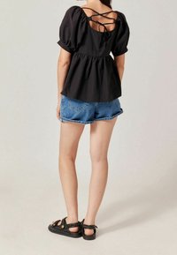 Black puff-sleeve top with a fitted bodice and gathered waist, featuring a crisscross back tie detail, paired with denim shorts and black sandals.