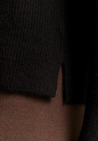 Schwarzer gerippter Strickpullover mit einem geschlitzten Saum, übereinander getragen mit einem braunen Stoff, der eine weiche Textur und unterschiedliche Farbtöne zeigt.