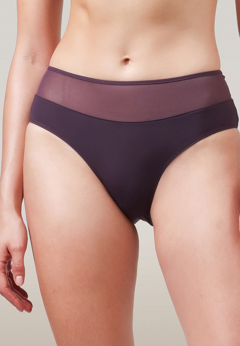 Kvinna iklädd högmidjade mörklila underkläder med en genomskinlig midja, stående mot en neutral bakgrund.