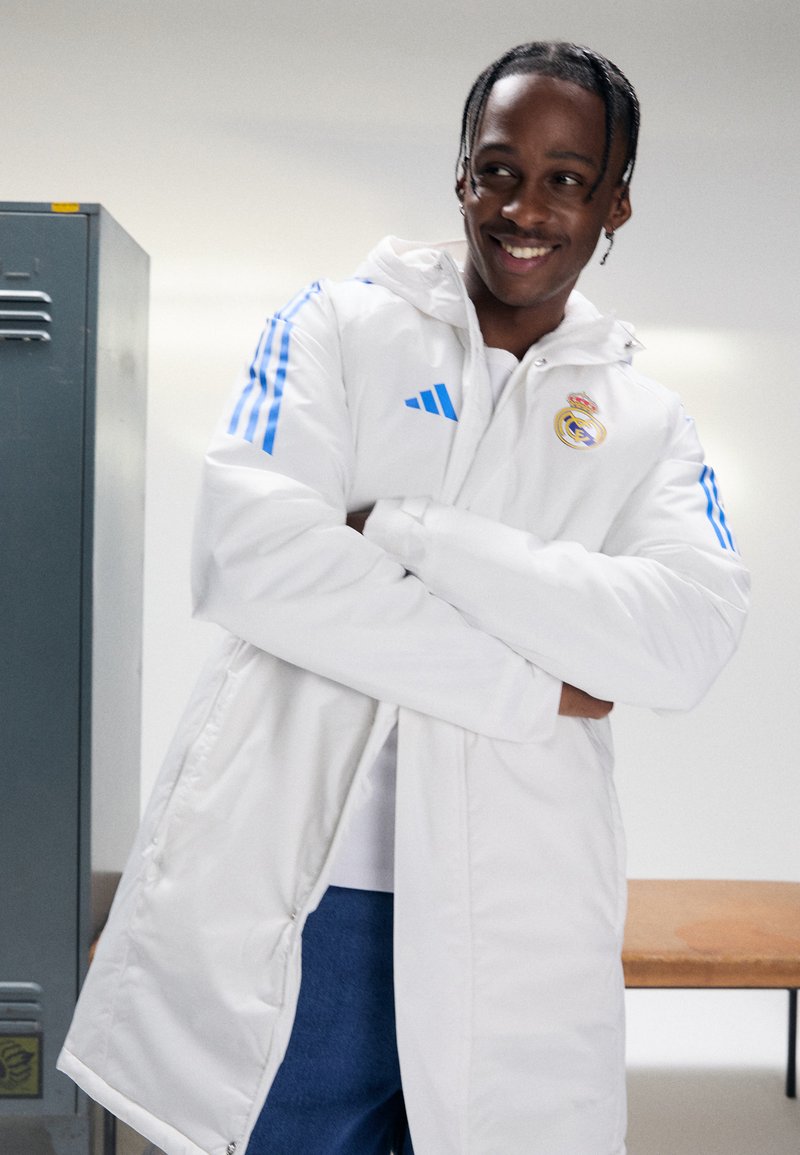 White hooded jacket with blue Adidas stripes and Real Madrid logo, featuring a smooth texture and a relaxed fit.