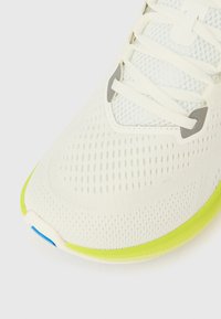 Κοντινή λήψη της μύτης ενός λευκού αθλητικού παπουτσιού από δίχτυ, με λευκά κορδόνια, γκρίζες λεπτομέρειες �στα οπές και κίτρινη σόλα με μια μικρή μπλε λεπτομέρεια στο μπροστινό μέρος.