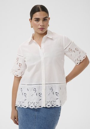 Woman with dark hair in a white button-up blouse featuring eyelet lace on sleeves and hem, paired with a blue denim skirt.