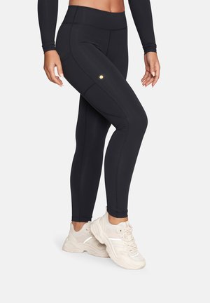 Kobieta w czarnych legginsach z wysokim stanem i detalami szwów oraz beżowych masywnych trampkach, stoi na jasnym, jednolitym tle.