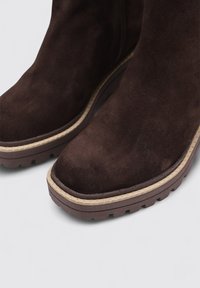 Botas de ante de color marrón oscuro hasta el tobillo con punta redonda y suela de goma de color crema. Presentan un perfil plano y una superficie texturizada.