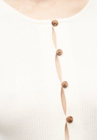 Κοντινό πλάνο ενός λευκού ριμπ υφάσματος με τέσσερα καφέ κουμπιά, μερικώς ξεκουμπωμένο, αποκαλύπτοντας το δέρμα κάτω από τη γραμμή των κουμπιών.