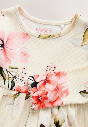 Lysecremefarvet kjole med ribstrikket stof, blomsterprint i lyserød og grøn, samt en flosset kantdetalje ved ærmekanten. Mærkeetiket synlig inde i halsudskæringen.