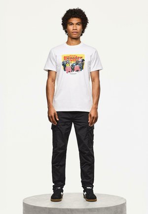 Vit t-shirt med en färgglad grafisk design, tillsammans med svarta cargo-byxor och svarta sneakers med vita detaljer.