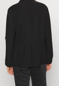 Veste noire à la coupe décontractée, avec une taille élastique et des manches longues. Présente un léger éclat, des détails de couture au dos et une poche latérale.