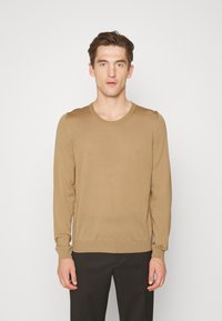 Pulover lung, de culoare maronie, cu maneci lungi, fabricat dintr-un material ușor, cu guler rotund și mansete cu țesătură dublă, purtat peste pantaloni închisi la culoare, pe un fundal simplu.