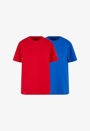 Jonge man met kort haar die een effen felblauw T-shirt met korte mouwen en een zwarte broek draagt, staat met zijn handen in zijn zakken.