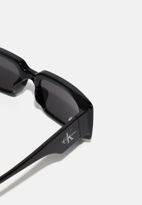 Fekete téglalap alakú napszemüveg sötét lencsékkel, fényes felülettel, az oldalsó szárakon ezüst Calvin Klein logóval. Szögletes és modern dizájn.