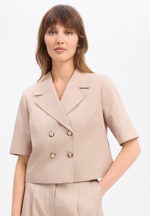 Vrouw met lichtbruin haar draagt een beige kort mouw dubbelgeknoopt blazer en bijpassende hoge taille broek, staat met één hand in de zak.
