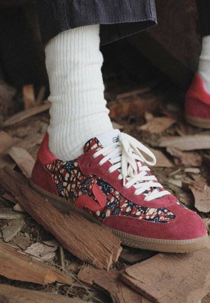 Pied portant une chaussette blanche côtelée et une basket rouge à motif floral avec des lacets blancs, marchant sur des débris de bois en plein air.