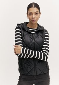 Chaleco negro acolchado con capucha, que presenta un cierre frontal y dos bolsillos laterales, usado sobre una camiseta de manga larga a rayas en blanco y negro.