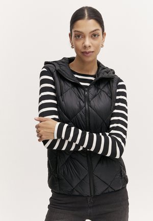 Zwarte gewatteerde vest met capuchon, voorzien van een ritssluiting aan de voorkant en twee zijzakken, gedragen over een zwart-wit gestreepte lange mouwen shirt.