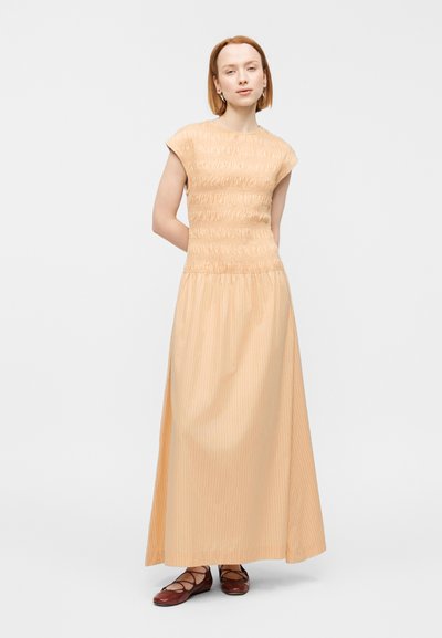 Μάξι φόρεμα από ελαφρύ ύφασμα σε ανοιχτή ροδακινί απόχρωση με σφηκοφωλιά στο πάνω μέρος και κοντά μανίκια, με φαρδύ, ριγέ κάθετα φούστα. Φορεμένο με καφέ επίπεδα παπούτσια.