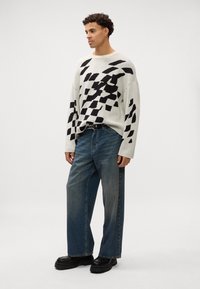 Strikkegenser i kremfarge med svart abstrakt mønster, parret med løse blå denimjeans og kraftige sorte platåer.