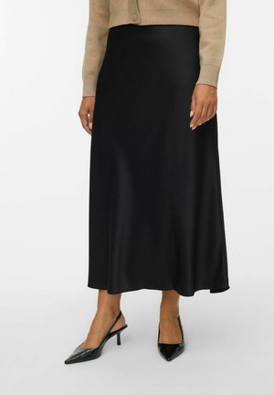 Frau trägt einen schwarzen Midirock, eine beige Strickjacke und schwarze spitze Slingback-Absätze und steht vor weißem Hintergrund.