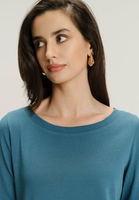 Kobieta z długimi ciemnymi włosami, nosząca złote kolczyki w kształcie łzy oraz niebieską bluzkę, patrząca w lewo na tle jasnego, jednolitego tła.