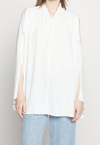 Camicia bianca con maniche aperte, lacci al collo, tessuto fluido e spacchi laterali. Abbinata a jeans in denim azzurro chiaro.