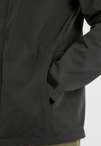 Black waterproof jacket with a smooth texture, featuring side pockets and a velcro accent near the cuff, showcasing a minimalist design.