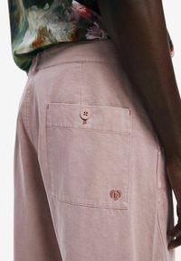 Helle rosa Hosen verfügen über eine Gesäßtasche mit Knopfverschluss und tonal abgesteppten Nähten. Ein Logo ist dezent in der Nähe der Tasche gestickt.