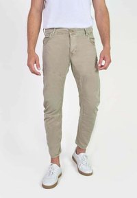 Man met beige tapered broek en witte sneakers, staande tegen een effen witte achtergrond.