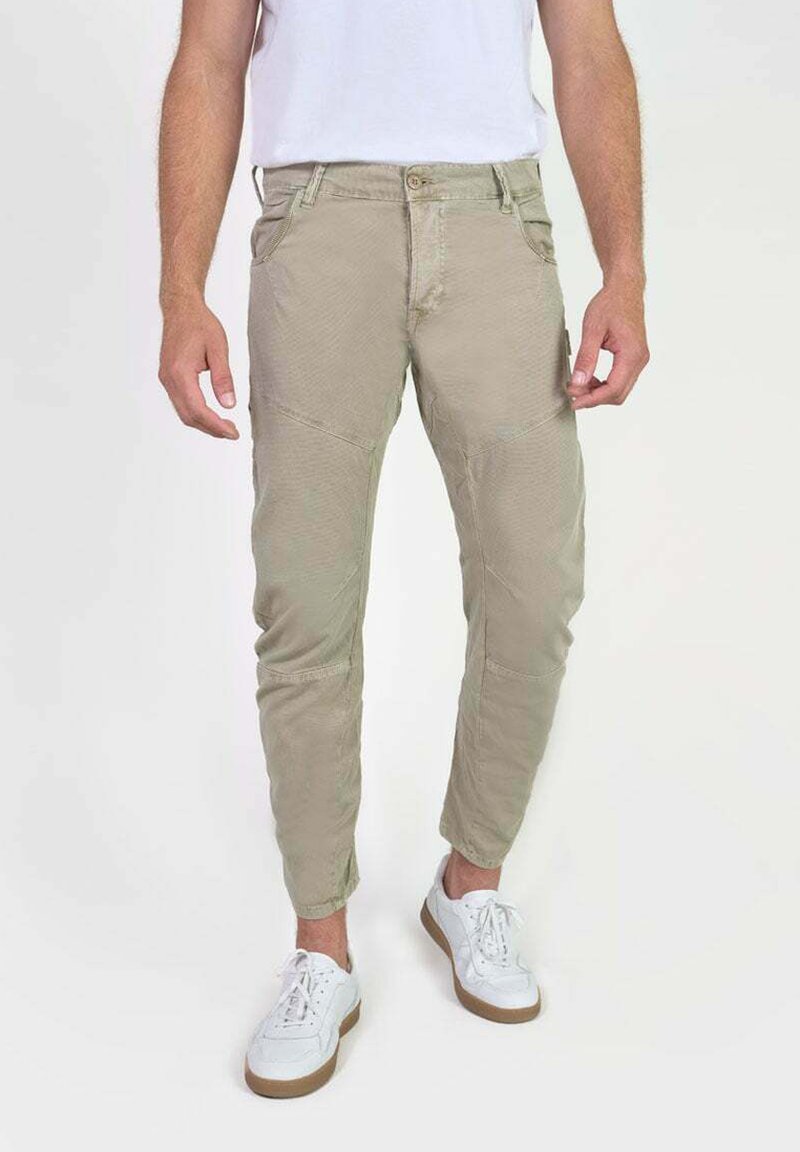 Man met beige tapered broek en witte sneakers, staande tegen een effen witte achtergrond.