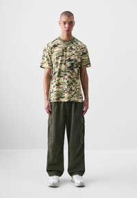 Camo-patterned short-sleeve shirt in beige, green, and blue, paired with loose olive cargo pants and white athletic shoes.