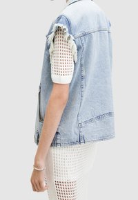Ljusa blå denimväst med en sliten ärmlös design, som har fransade kanter och en dragkedja fram. Bärs över en vit nättröja.
