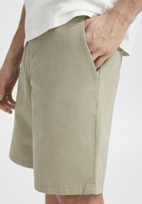 Khaki värvi lühikesed püksid, mis on valmistatud tekstuuriga kangast, külje taskute ja vabalt lõigatud disainiga. Ahned servad annavad puhta viimistluse.