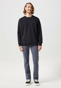 Felpa nera con scollatura a girocollo, polsini a costine e un piccolo logo sul petto, abbinata a pantaloni grigi e sneakers nere.