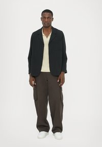 Schwarze Blazer mit einer Brusttasche, kombiniert mit einem hellbeigen Polohemd und dunklen Cargo-Hosen. Weiße Sneaker runden das Outfit ab.