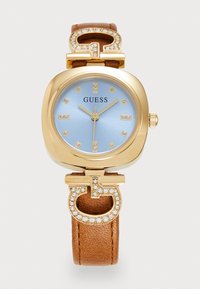 Goldfarbene Guess-Armbanduhr mit hellblauem Ziffernblatt, Kristall-Stundenmarkierungen, mit Kristallen verzierten "G"-Akzenten und einem strukturierten braunen Lederarmband.