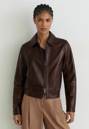 Frau mit geflochtenem Haar, die eine dunkelbraune Lederjacke und hellbraune weit geschnittene Hose trägt, steht vor einer schlichten weißen Wand.