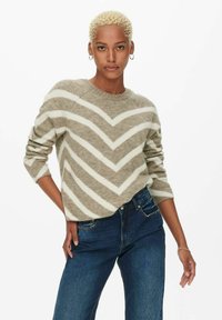 Pull rayé beige et crème avec un motif en chevrons, col rond et manches longues, associé à un jean bleu foncé. Matière en maille texturée.