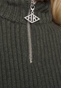 Maglione verde a coste con colletto alto e una cerniera argentata con un ciondolo a forma di diamante che mostra "HK" vicino al cursore.