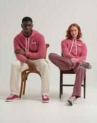 Felpe rosa con stringhe bianche e logo, pantaloni crema e sneakers rosse. Pantaloni a righe su figura seduta, entrambi su sedie di legno.