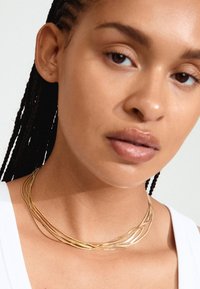 Guldtonad flerlagerhalsband med släta, böjda trådar. Bärs runt halsen och kompletterar en vit topp. Subtil glans och lätt design.