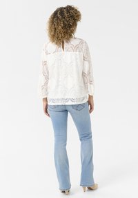 Blusa in pizzo bianco con maniche a tre quarti, caratterizzata da un motivo floreale e dettaglio a chiave sul retro, abbinata a jeans svasati di colore azzurro chiaro.