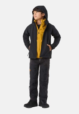 Junge Person steht mit den Händen in den Taschen, trägt eine schwarze Kapuzenjacke, ein gelbes Fleecepullover, schwarze Hose und schwarze Wanderstiefel, schaut nach links.