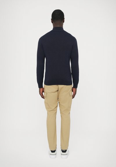 Camisola de malha canelada azul-marinho combinada com calças cargo bege. As calças têm bolsos laterais e são usadas com ténis brancos. Vista traseira.